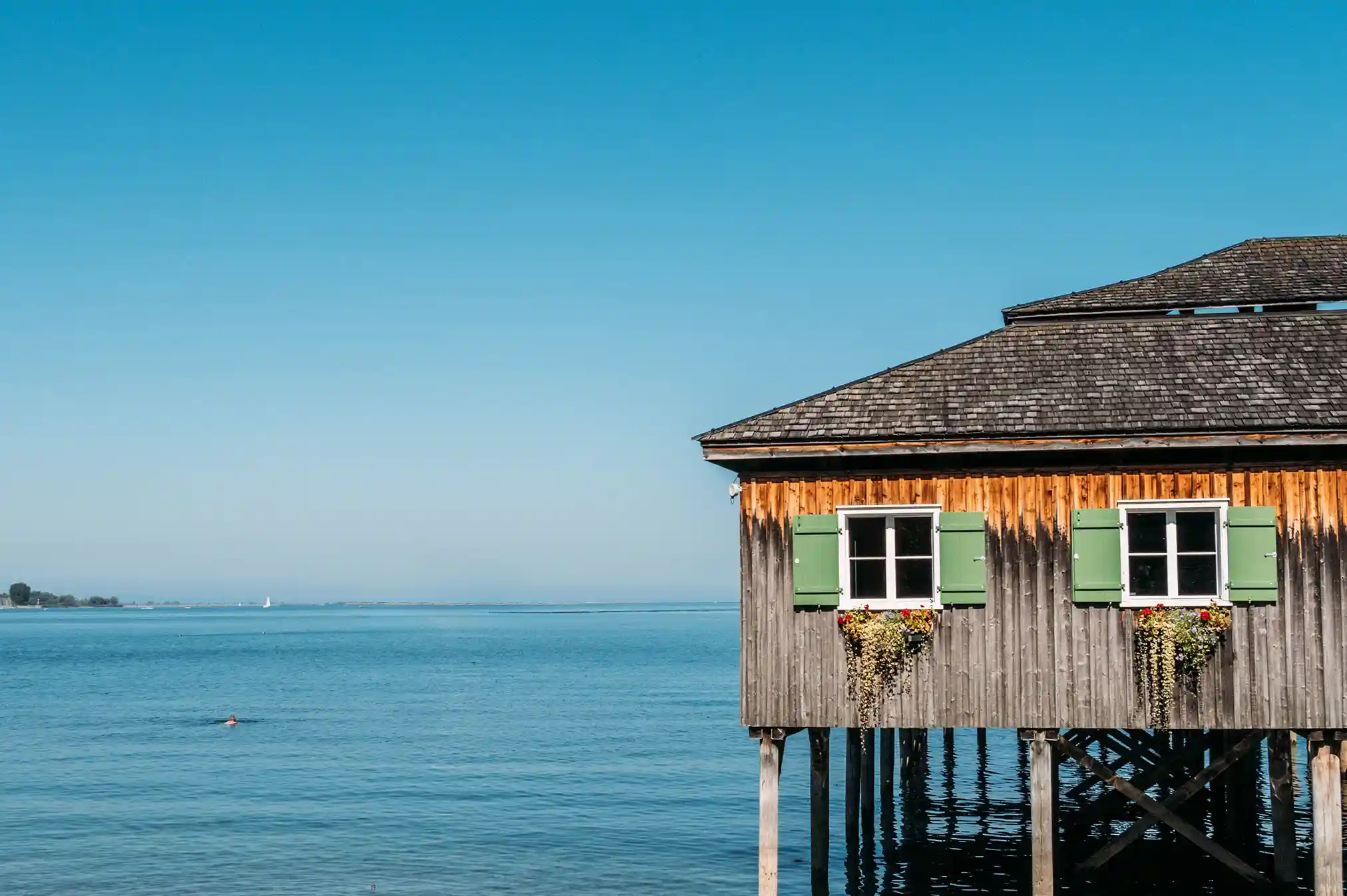 Das Holzhaus Mili auf Stelzen mit grünen Fensterläden und Person am Eingang und im Hintergrund der Bodensee