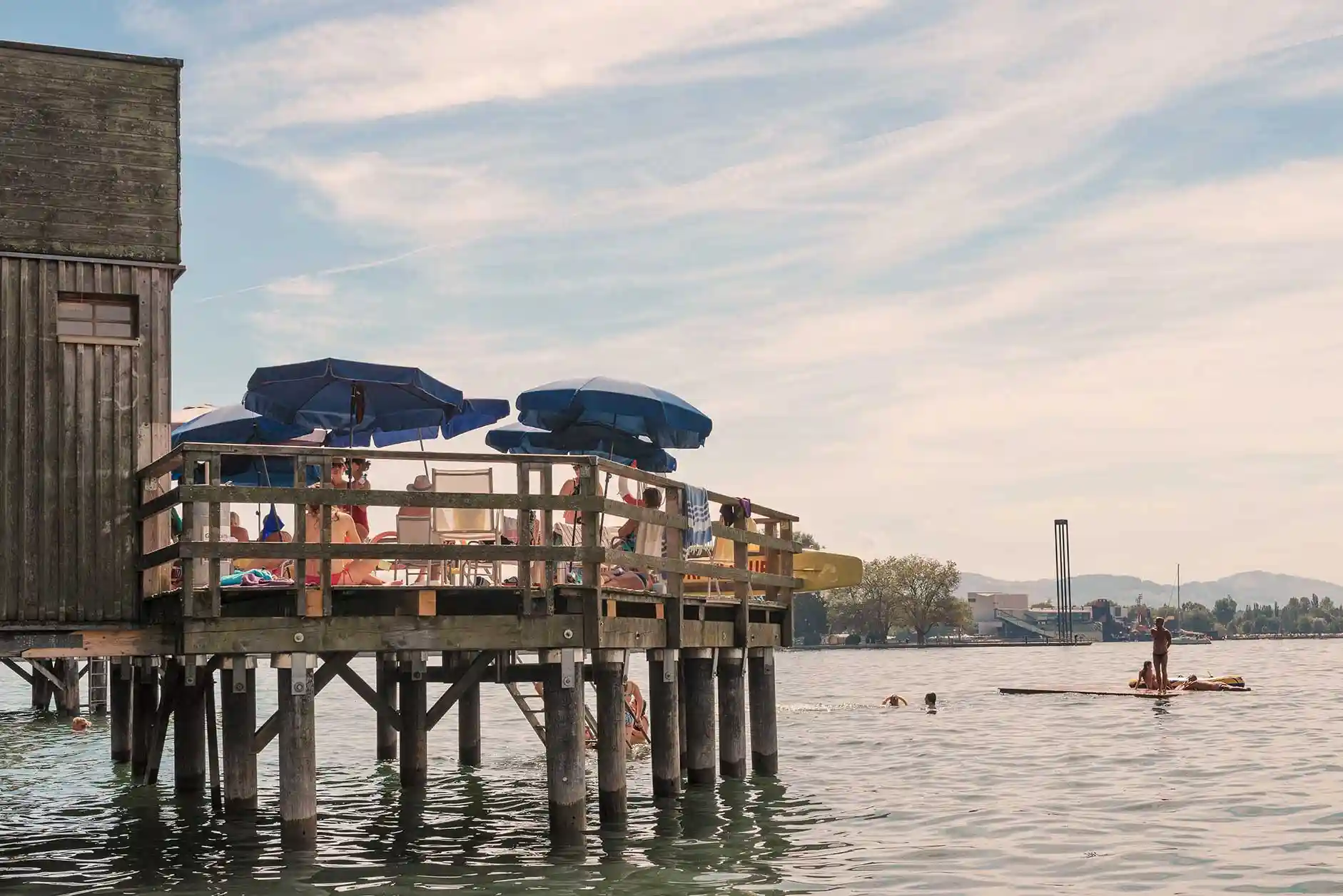 Menschen sitzen und liegen auf der Holzplattform der Mili am See, einige schwimmen im Wasser